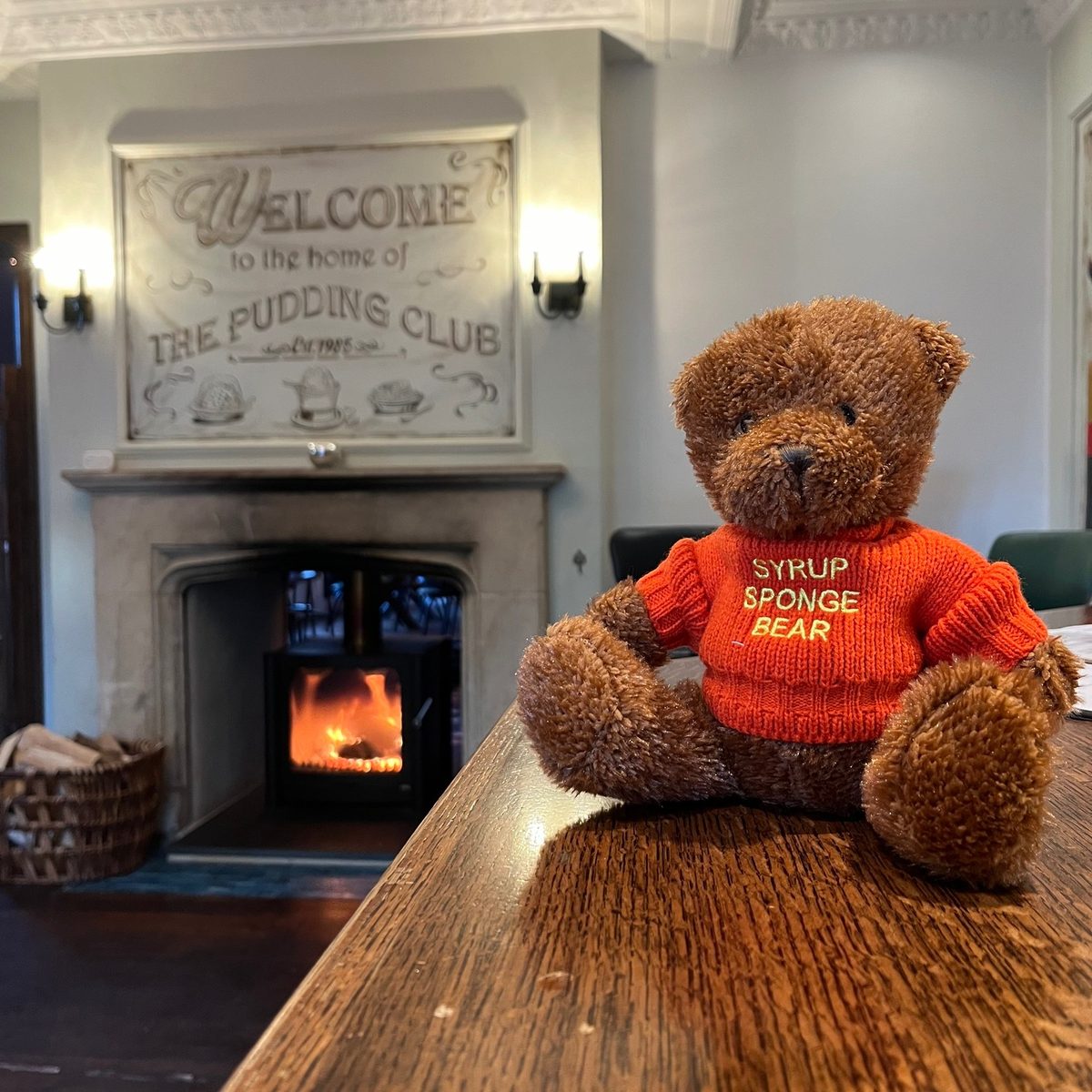Syrup Sponge Bear in a knitted jumper beside the log fire under the Welcome to the home of The Pudding Club sign